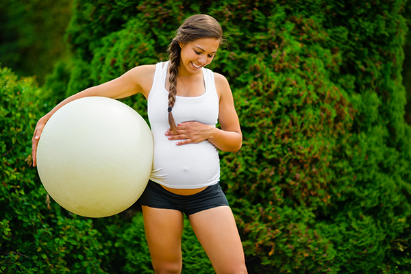 Smiling expectant mother touching stomach while holding exercise ball against trees in park