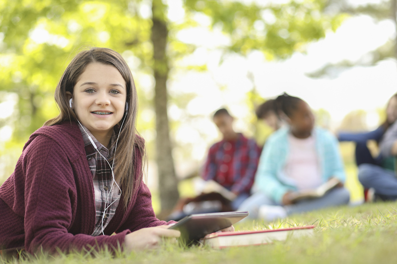 A middle school student enjoys the flexibility of online learning- tablet in hand, lying on green grass with other students in the background.