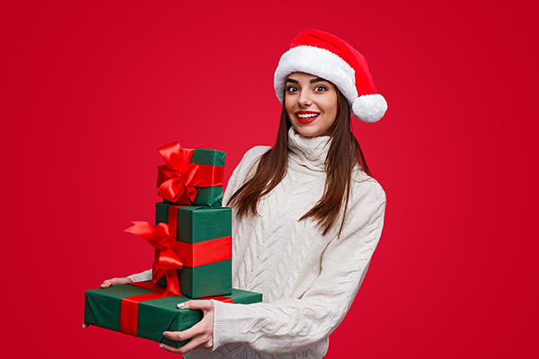 Happy young lady in Santa hat and sweater smiling and carrying stack of gift boxes in green wrapping on Christmas Day against red background