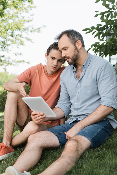 bearded dad and teenager son looking at digital tablet while sitting in green park