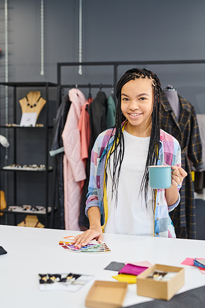 Portrait of young tailor with African braids standing and drinking coffee while working with colorful samples in atelier