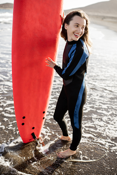 Portrait of a young woman in wetsuit standing with red surfboard on the ocean beach during a sunset. Water sport and active lifestyle concept