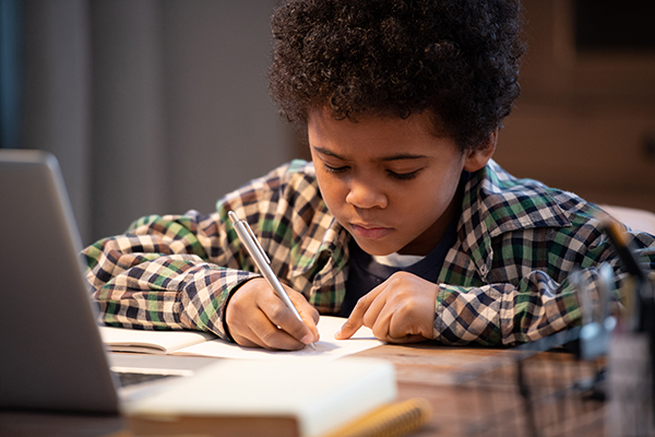Diligent little African schoolboy in casualwear making notes in copybook while sitting in front of laptop and carrying out home assignment