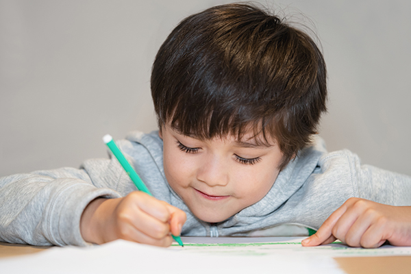 Candid shot  happy kid sitting alone doing homework, Child boy with smiking face holding green pen drawing and colouring on white paper, Elementary school and homeschooling concept